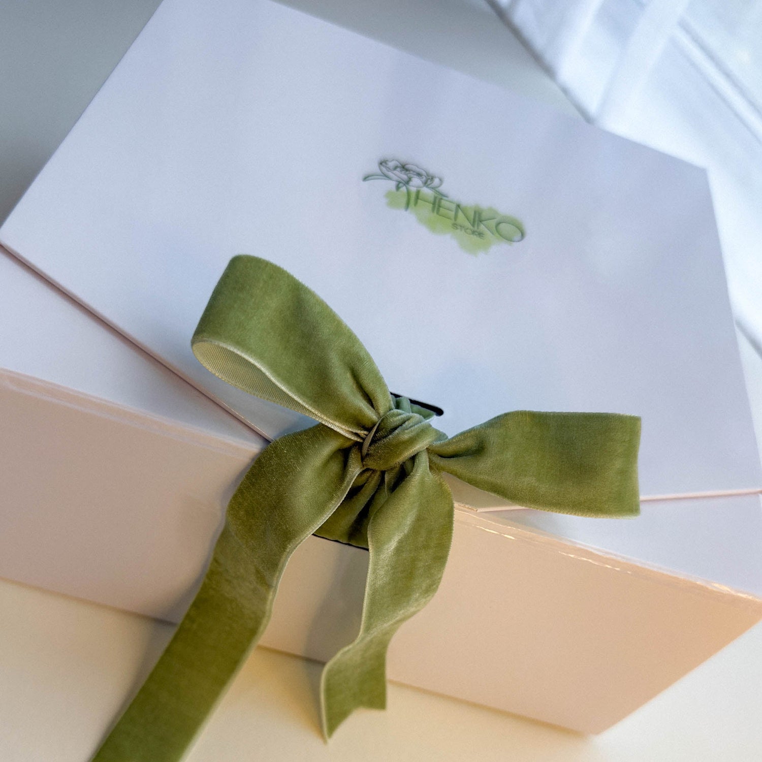 Cream gift box tied with a sage green satin ribbon bow, surrounded by candles, skincare products and soft linen fabric. Part of Henko Store's The Bloom Hour Mother's Day collection.