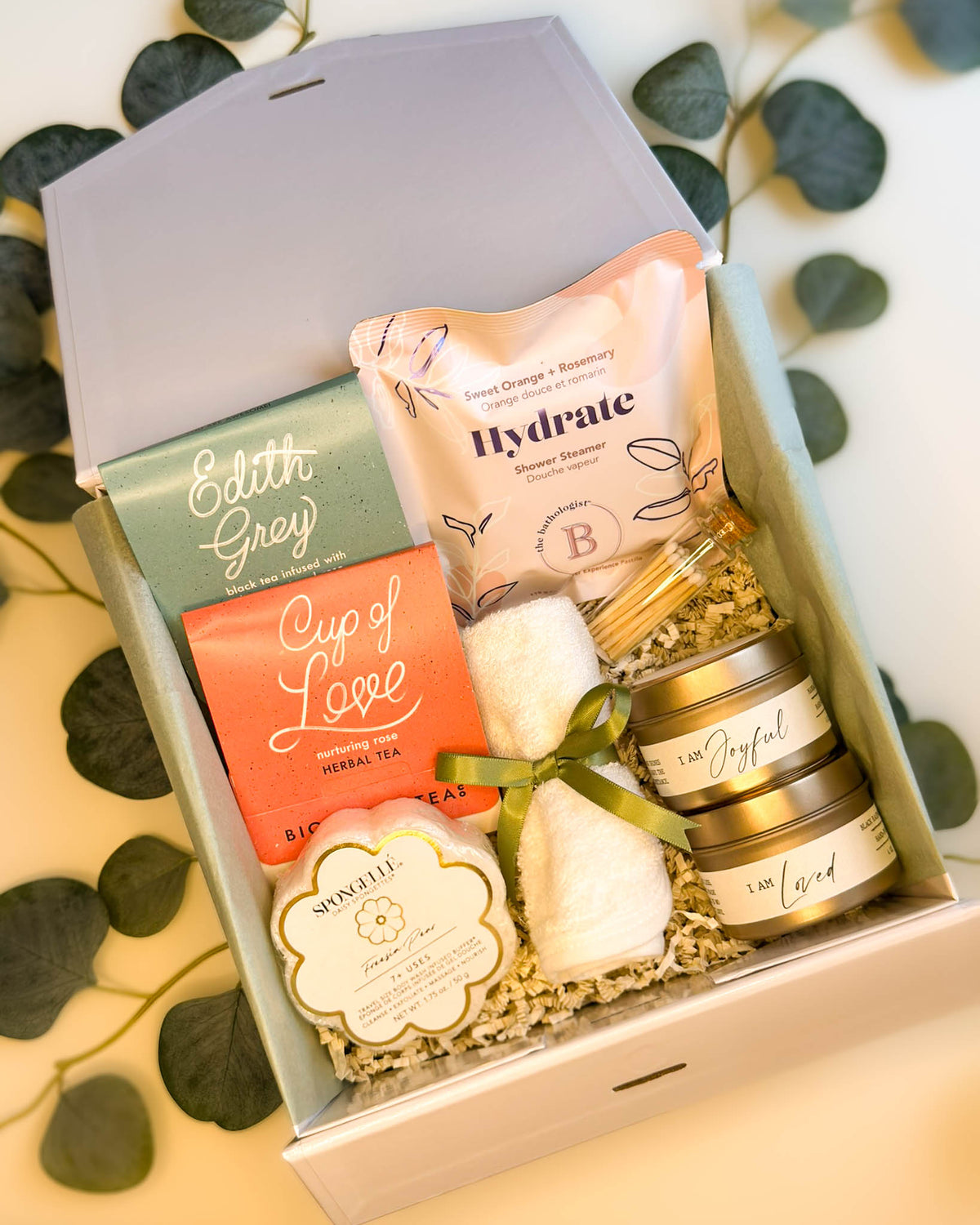 Gift box with various items including a tea bag, shower steamer, sponge, and candle on a light background.